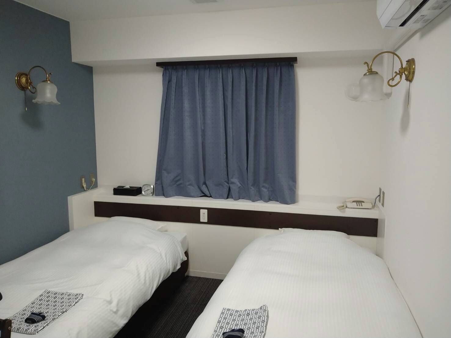 A hotel room with two single beds, a window with blue curtains, a bedside table with a phone, and wall-mounted light fixtures.