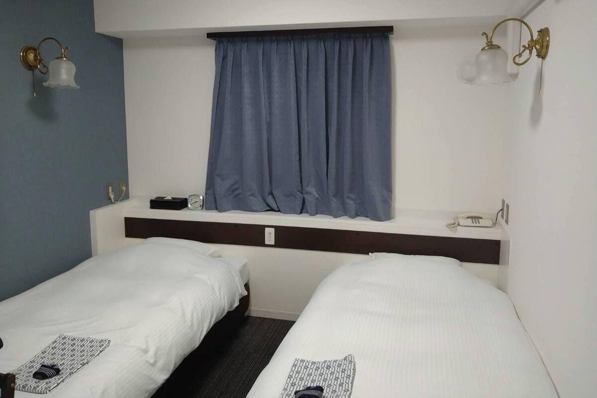 A hotel room with two single beds, a window with blue curtains, a bedside table with a phone, and wall-mounted light fixtures.