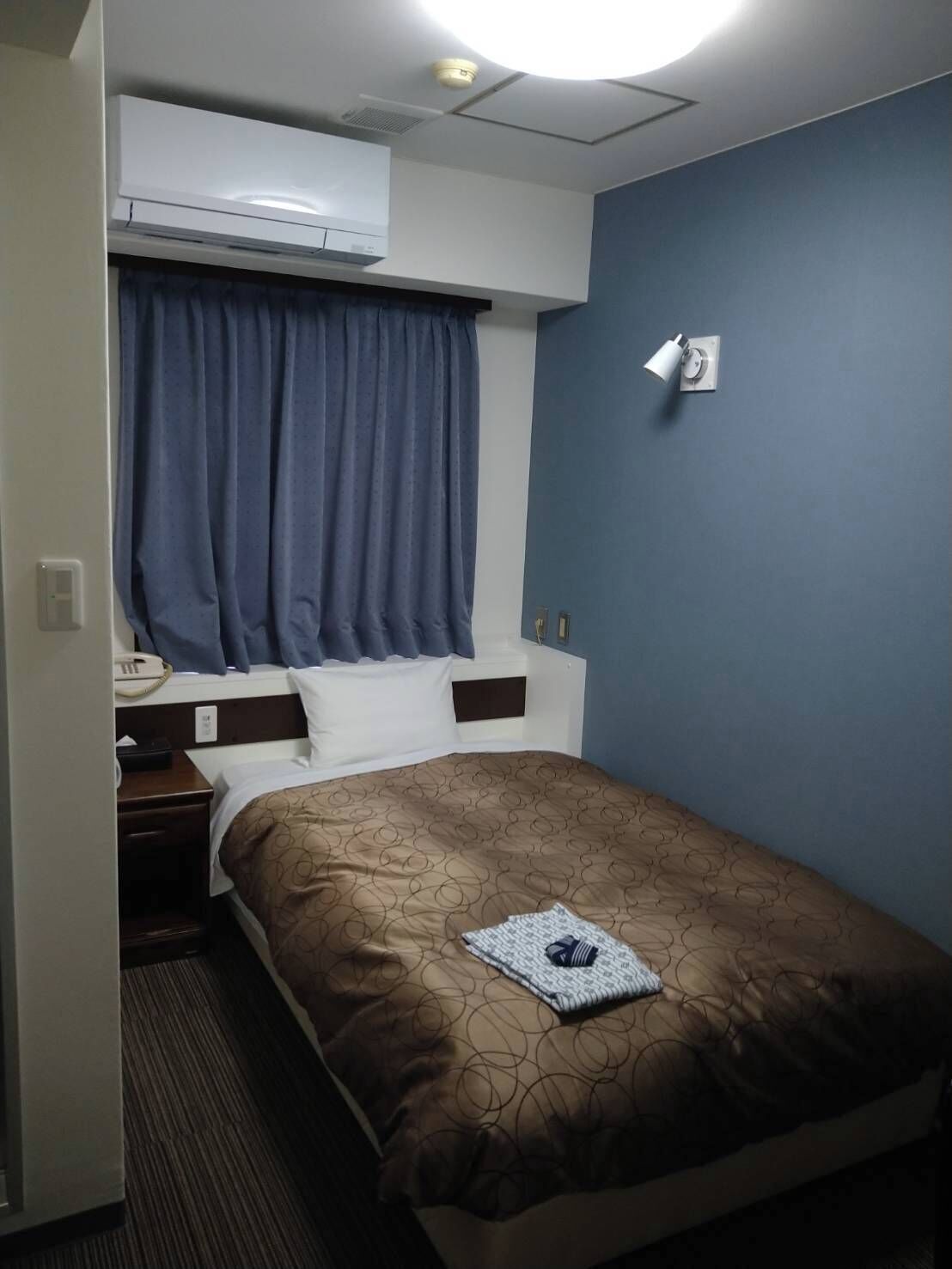 A hotel room with a bed, a nightstand, a telephone, an air conditioning unit, a window with curtains, and a wall-mounted light fixture.