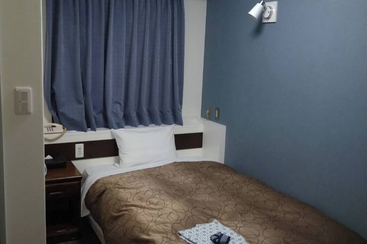 A hotel room with a bed, a nightstand, a telephone, an air conditioning unit, a window with curtains, and a wall-mounted light fixture.