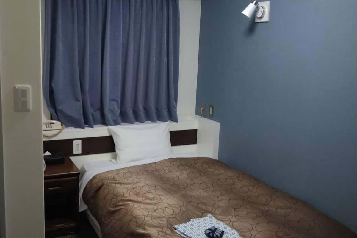 Une chambre d’hôtel comprenant un lit, une table de chevet, un téléphone, un appareil de climatisation, une fenêtre avec des rideaux et un luminaire fixé au mur.