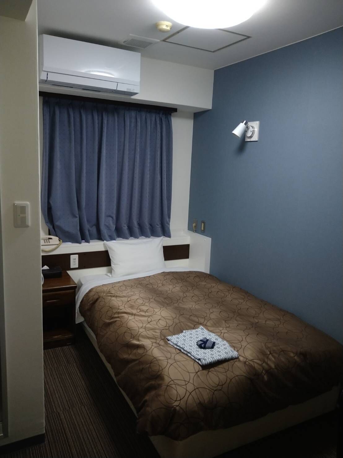 A hotel room with a bed, a bedside table, a phone, a wall-mounted light, and an air conditioning unit.