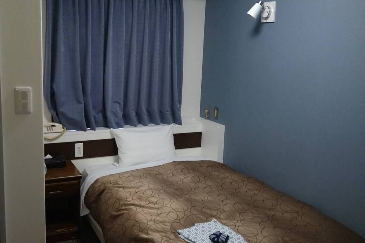 A hotel room with a bed, a bedside table, a phone, a wall-mounted light, and an air conditioning unit.