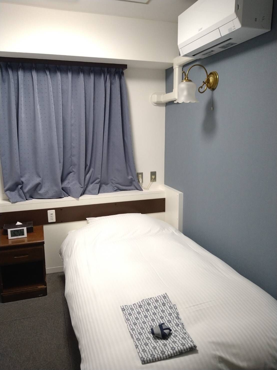 A hotel room with a bed, a nightstand, a window with curtains, a wall-mounted air conditioner, and a bedside lamp.