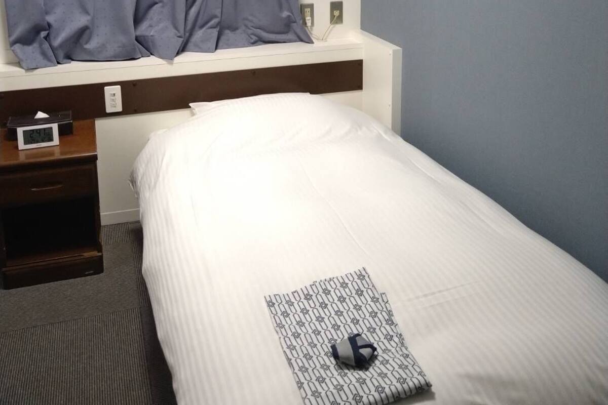 A hotel room with a bed, a nightstand, a window with curtains, a wall-mounted air conditioner, and a bedside lamp.