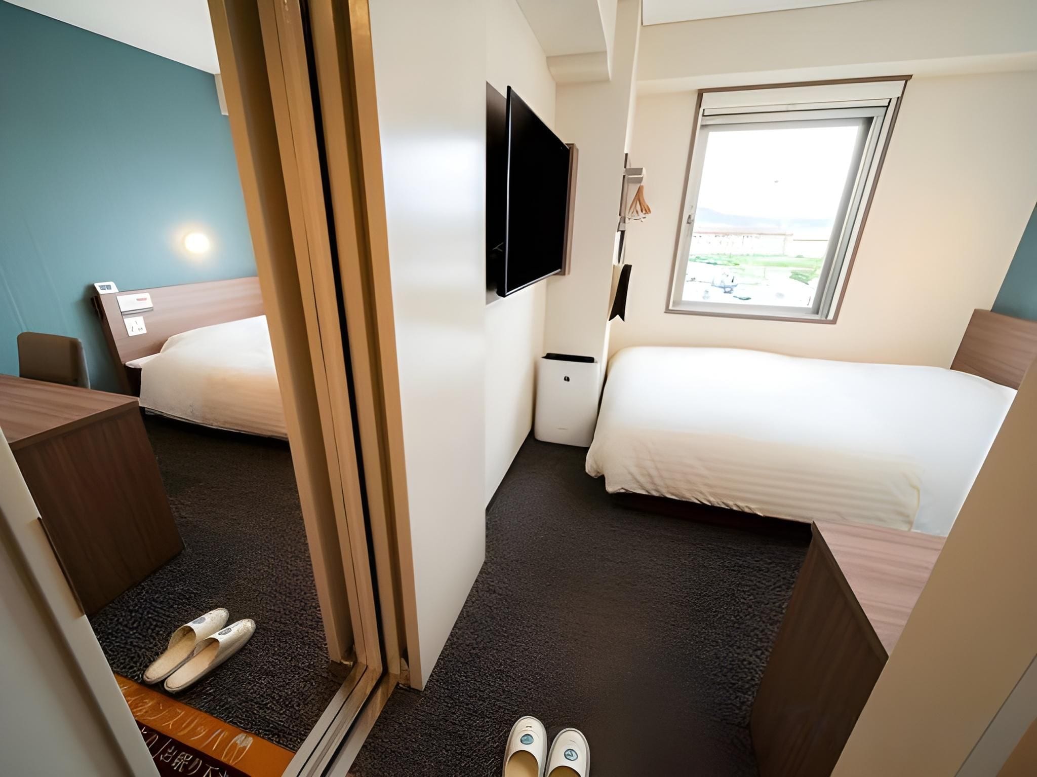 A compact hotel room with two beds, a desk, a TV, and a window with a view.