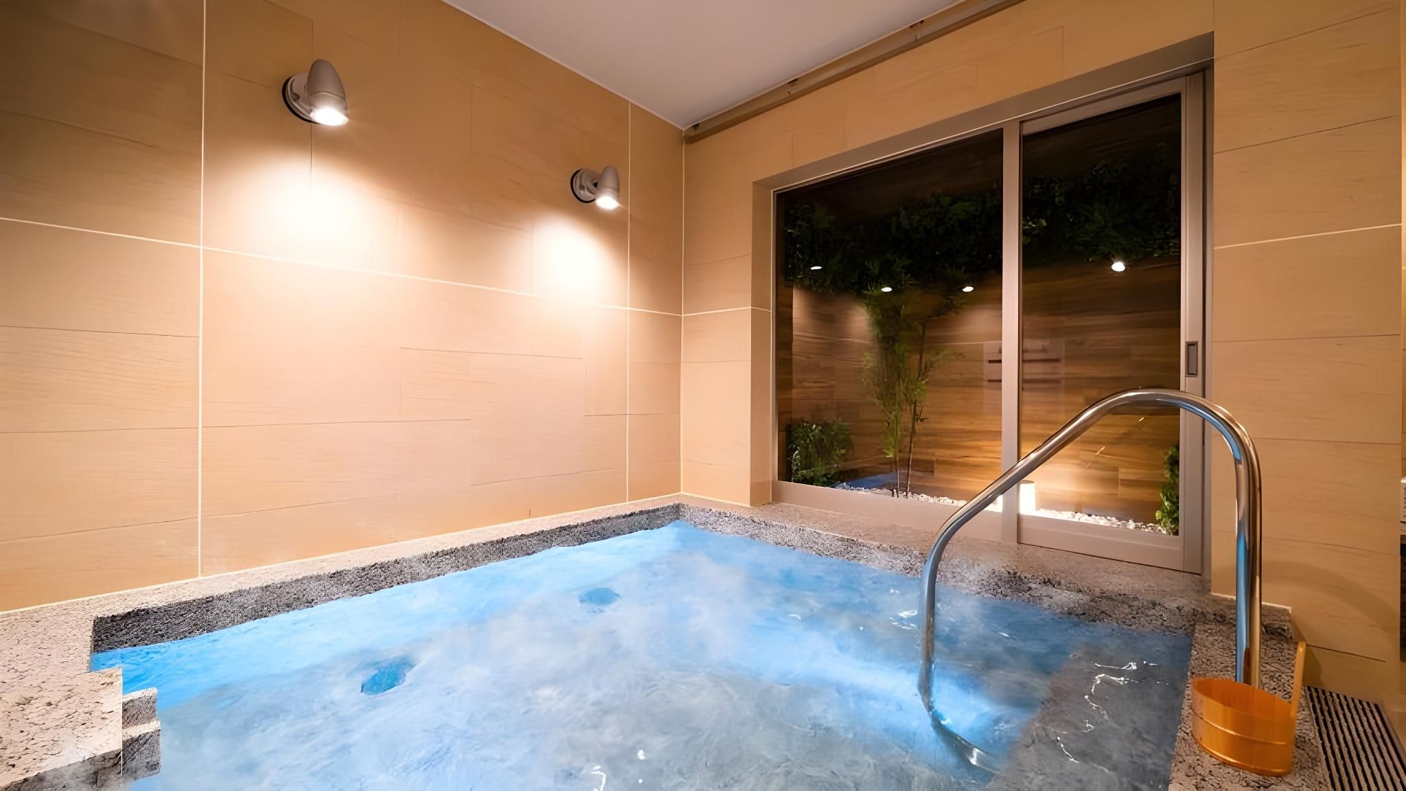 A spa area with a hot tub, a metal handrail, and a view of greenery outside.