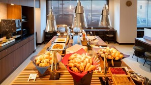Montaje de bufé con diversos alimentos, incluyendo pan y pastelería, en un moderno hotel con suelo de madera y ventanas amplias.