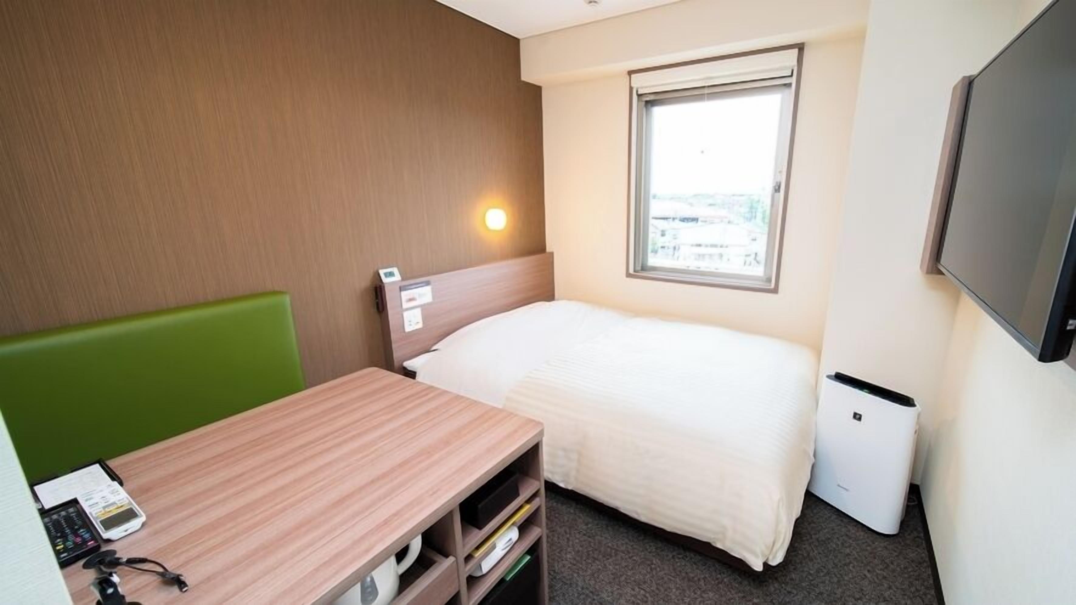 Una habitación de hotel pequeña con una cama individual, un escritorio con silla, una mesita, un televisor y una ventana con vistas a edificios.