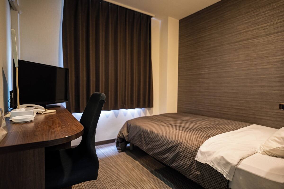 Una habitación de hotel con cama, escritorio, silla, televisión y ventana con cortinas.