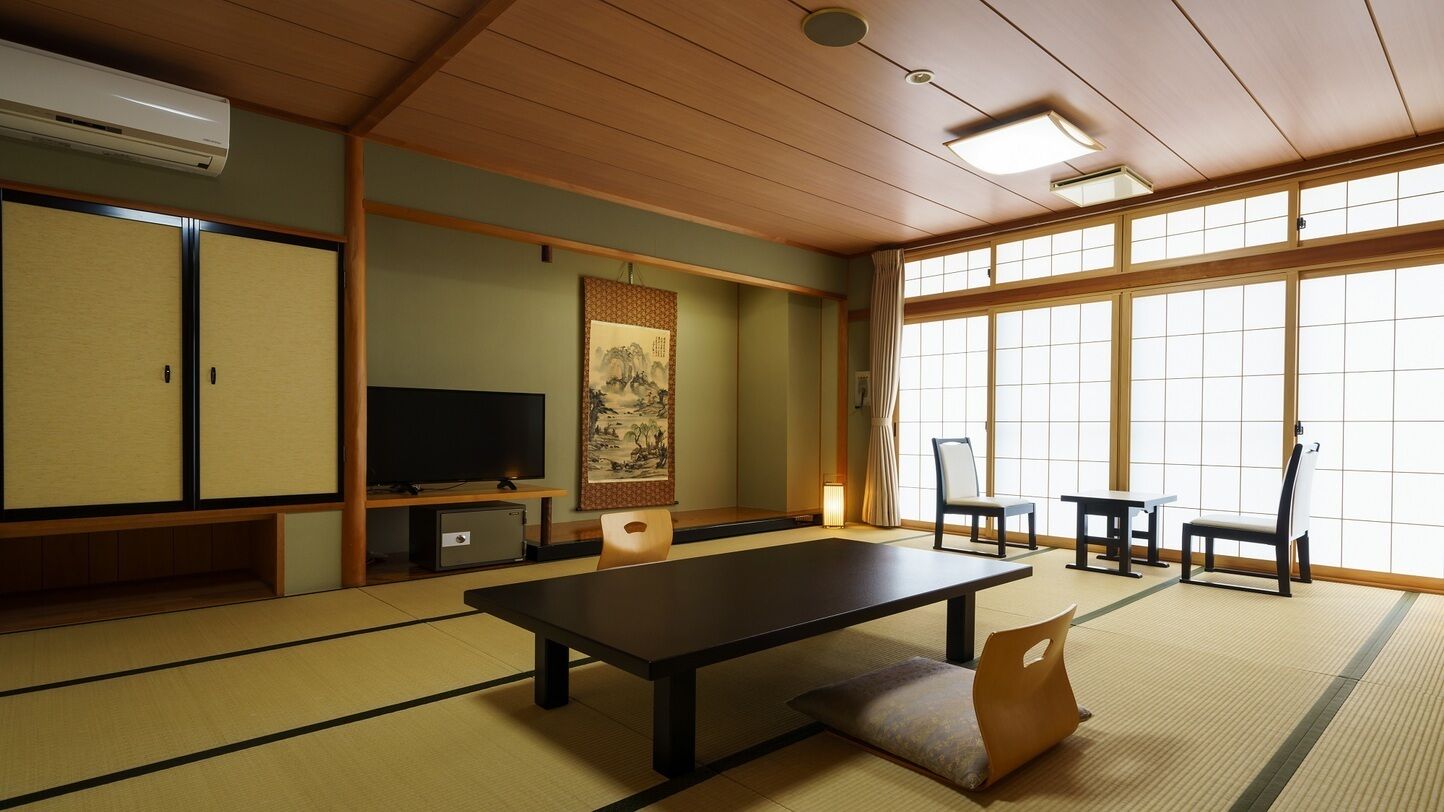 Ein traditioneller japanischer Raum mit Tatami-Bodenbelag, einem niedrigen Tisch, Stühlen, einem Fernseher und Schiebetüren.