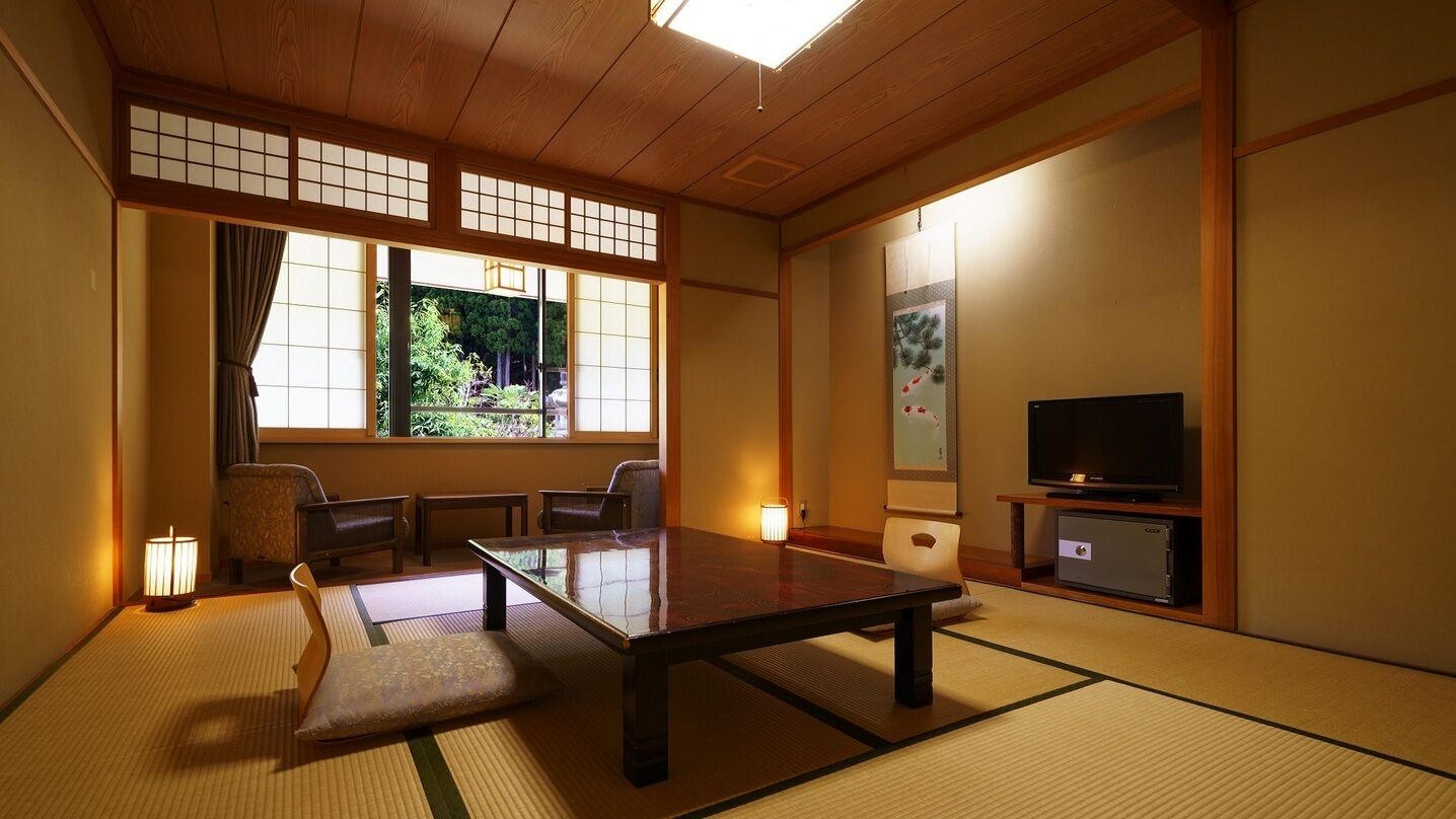 Ein traditioneller japanischer Raum mit Tatami-Boden, einem niedrigen Tisch, Sitzgelegenheiten, einem Fernseher und einer Schiebetür.