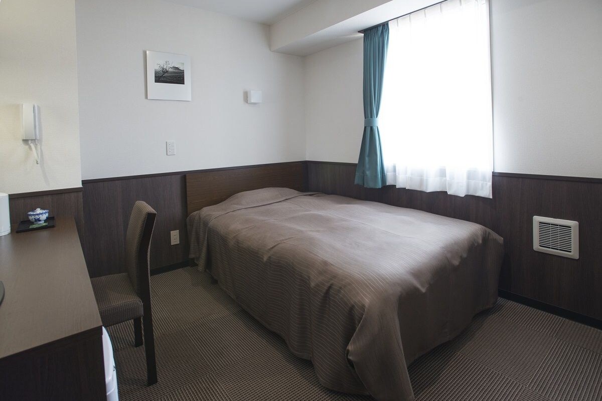 A single bed with a headboard, a desk with a chair, a window with curtains, and a wall-mounted light.