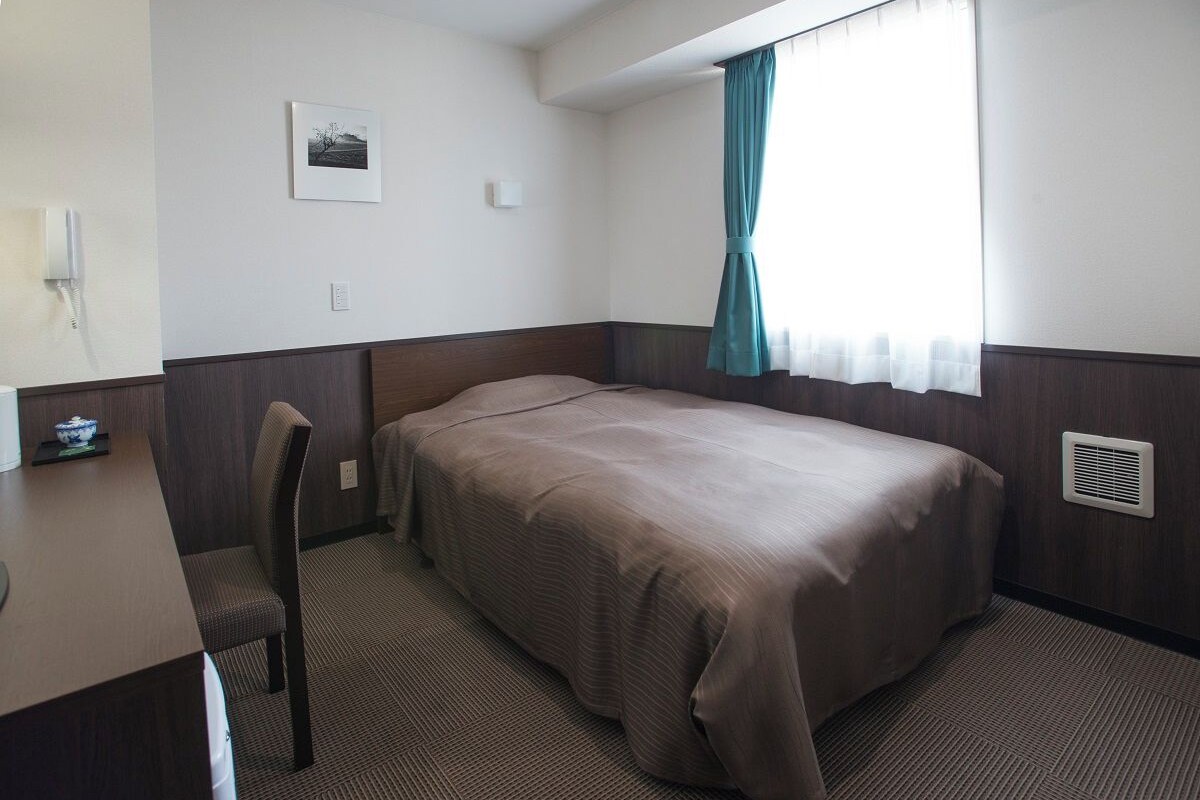 A single bed with a brown bedspread, a chair, a desk with a small table, a window with a curtain, and a picture on the wall.