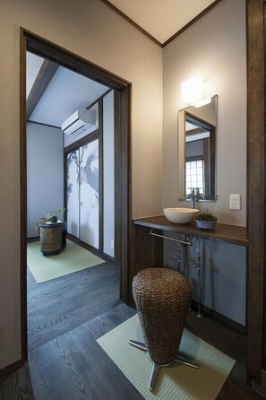 A hallway with a wooden floor, a wicker chair, a sink with a mirror, and a plant on the counter. - Daimaru An (Kanazawa)