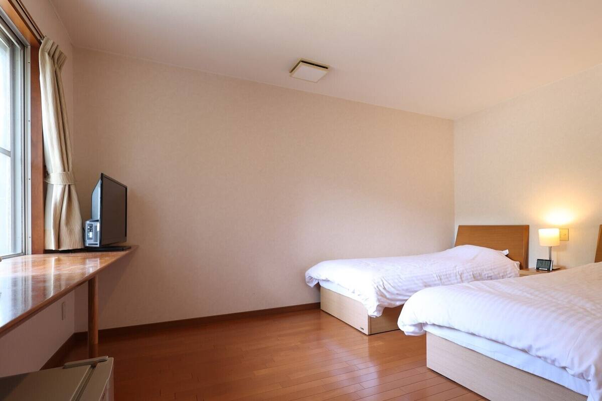 Ein Hotelzimmer mit zwei Betten, einem Schreibtisch mit Computer, einem Fenster mit Vorhängen und einer Wandleuchte.