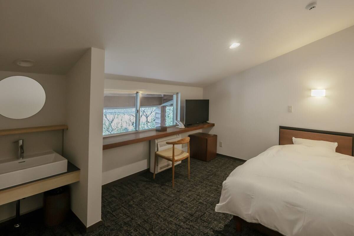 Ein Hotelzimmer mit einem Bett, einem Schreibtisch mit Stuhl, einem Fernseher, einem runden Spiegel und einem Fenster.