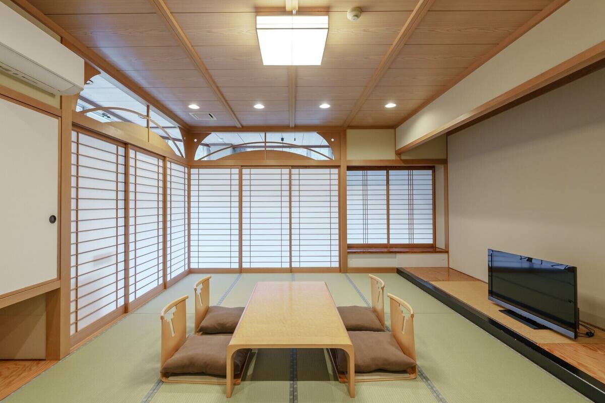 Ein traditioneller japanischer Raum mit Tatami-Boden, einem niedrigen Tisch mit Kissen, Schiebetüren und einem Fernseher.