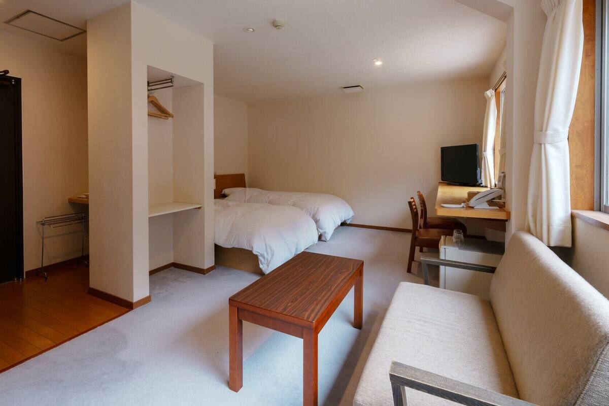 Ein gemütlicher Raum mit einem Bett, einem Holztisch, einer Couch, einem Schreibtisch mit Computer und einem Fernseher.