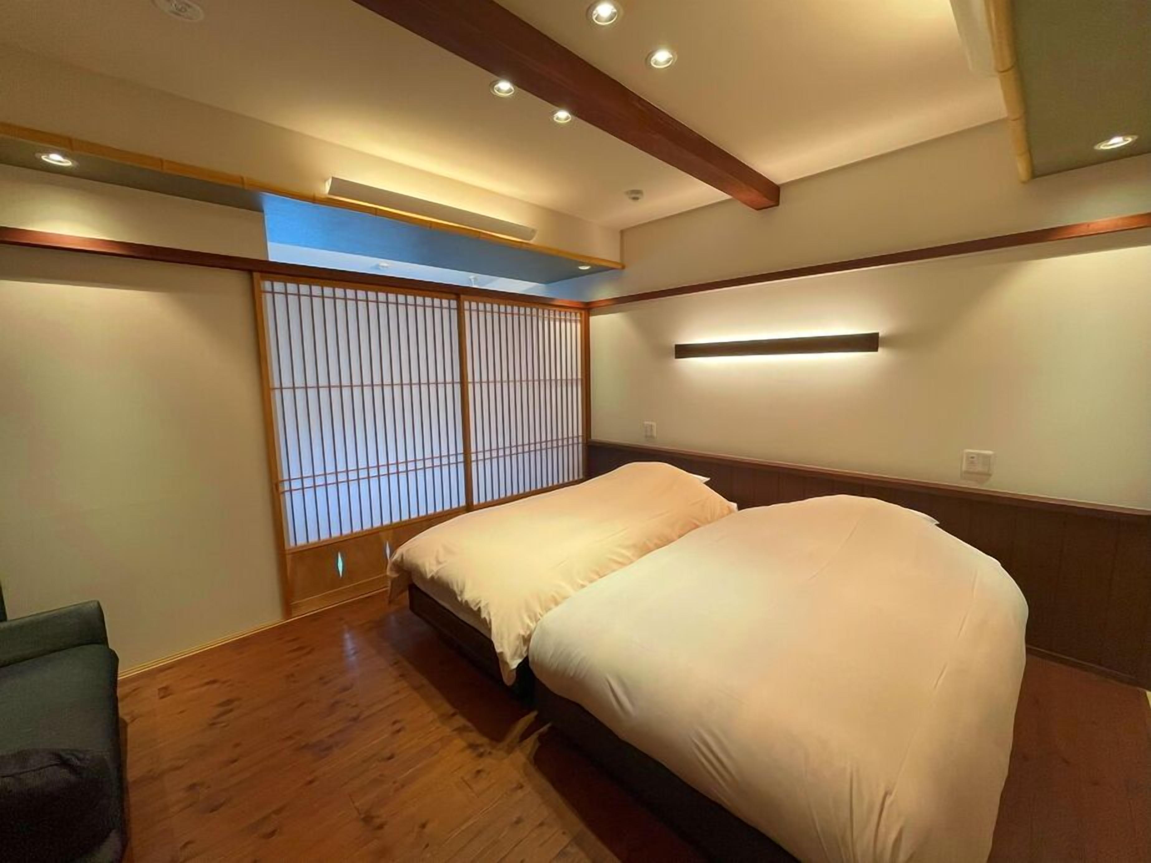 A room with a wooden floor, two beds with white bedding, a shoji screen, and recessed ceiling lighting.