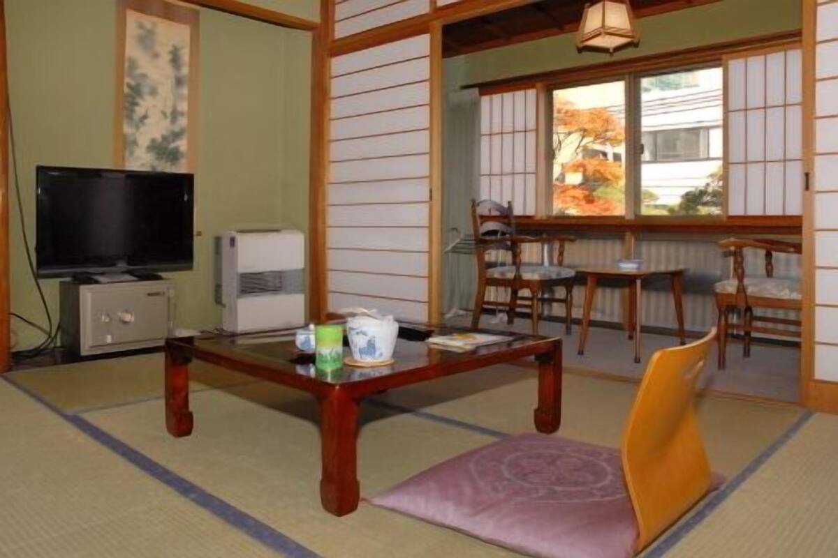 Um quarto tradicional japonês com tatames, televisão, pequeno frigorífico, mesa com almofada e vista para um jardim através de portas deslizantes.