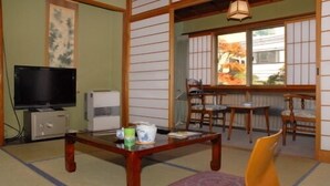 Perinteinen japanilaistyylinen huone, jossa on tatamimatto, televisio, pieni jääkaappi, tyynyillä varustettu pöytä ja liukuovista avautuva näkymä puutarhaan.