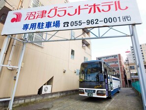 Un autobús estacionado bajo un letrero con texto japonés, un edificio de fachada color crema y un autobús azul con 'GALA' en la parte frontal.