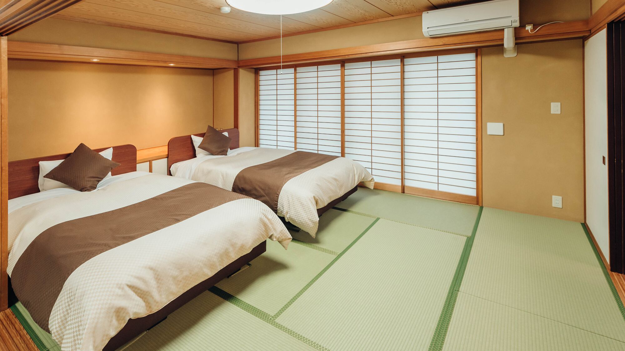 Ein traditioneller japanischer Raum mit Tatami-Boden, zwei Betten mit brauner Bettwäsche, einem verschiebbaren Shoji-Bildschirm und einer Klimaanlage.