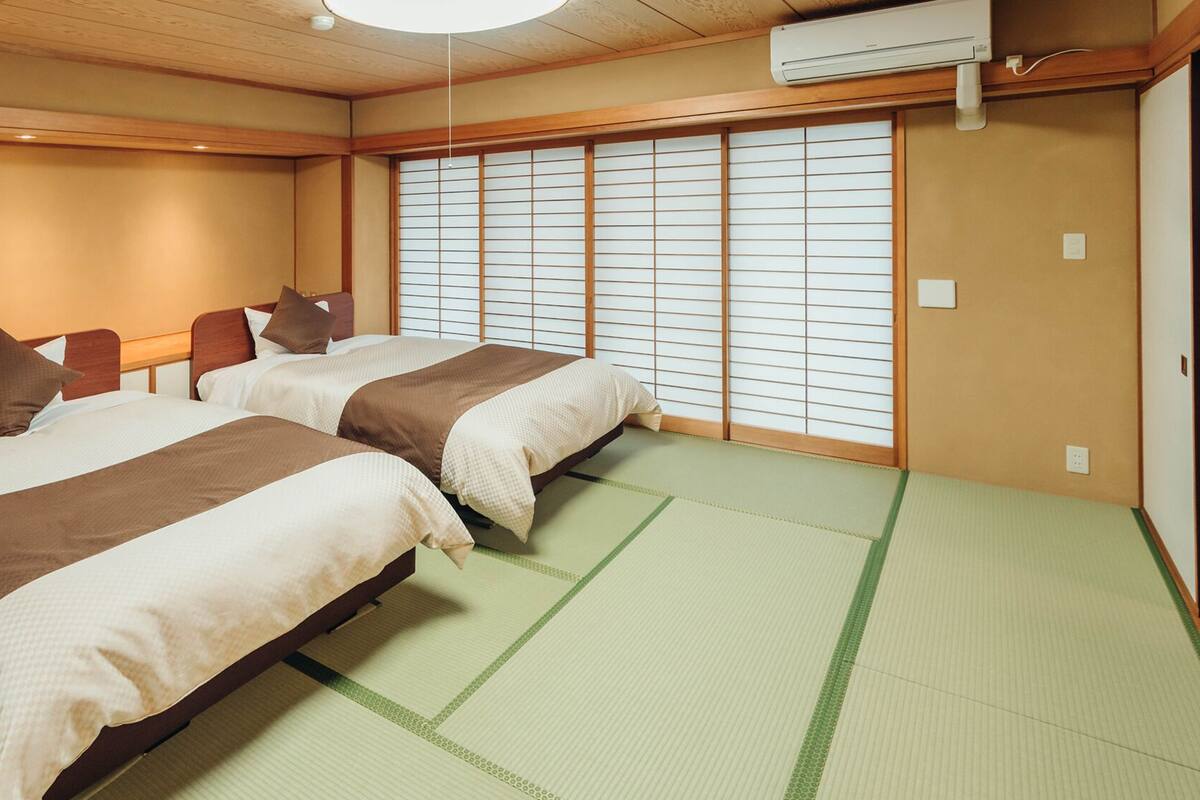 Una tradizionale stanza giapponese con pavimento in tatami, due letti con biancheria marrone, un paravento scorrevole shoji e un condizionatore d'aria.