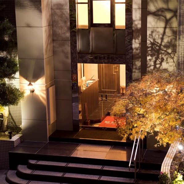 Ein Hotel mit Eingangsbereich und einer Treppe zum Eingang, einem kleinen Garten mit Baum sowie einer beleuchteten Außenwand.