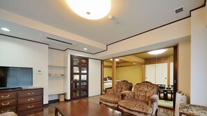 A spacious living room with a wooden coffee table, two patterned armchairs, a sofa, and a television on a wooden cabinet. - Hotel Showa Club (Atami)