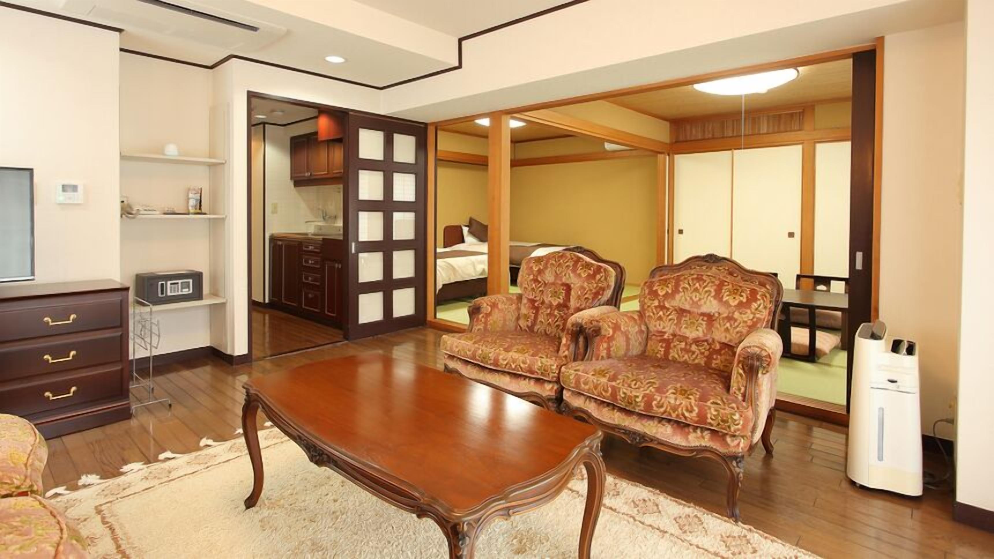 A traditional Japanese-style room with tatami flooring, a low wooden table, two patterned armchairs, a television, and a sliding door leading to a bedroom.