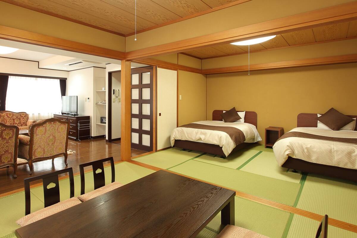 Una camera d'hotel in stile tradizionale giapponese, con pavimento in tatami, un letto basso in legno, una poltrona e un tavolo da pranzo.