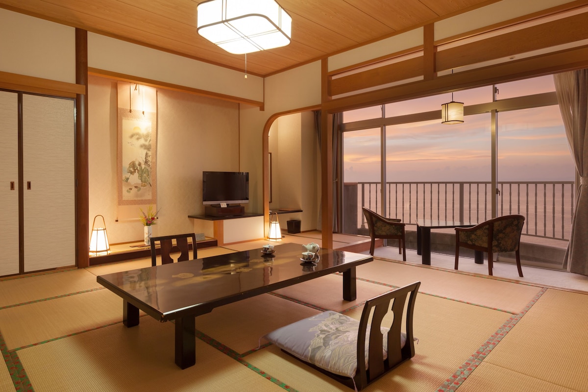 A traditional Japanese room with tatami flooring, a low table, chairs, a television, and a view of the sea.
