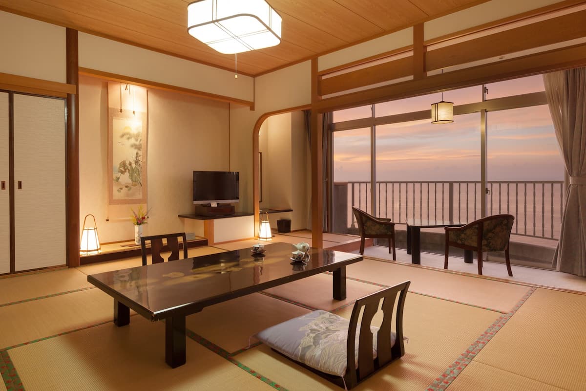 Una habitación tradicional japonesa con suelo de tatami, una mesa baja, sillas, un televisor y vista al mar.