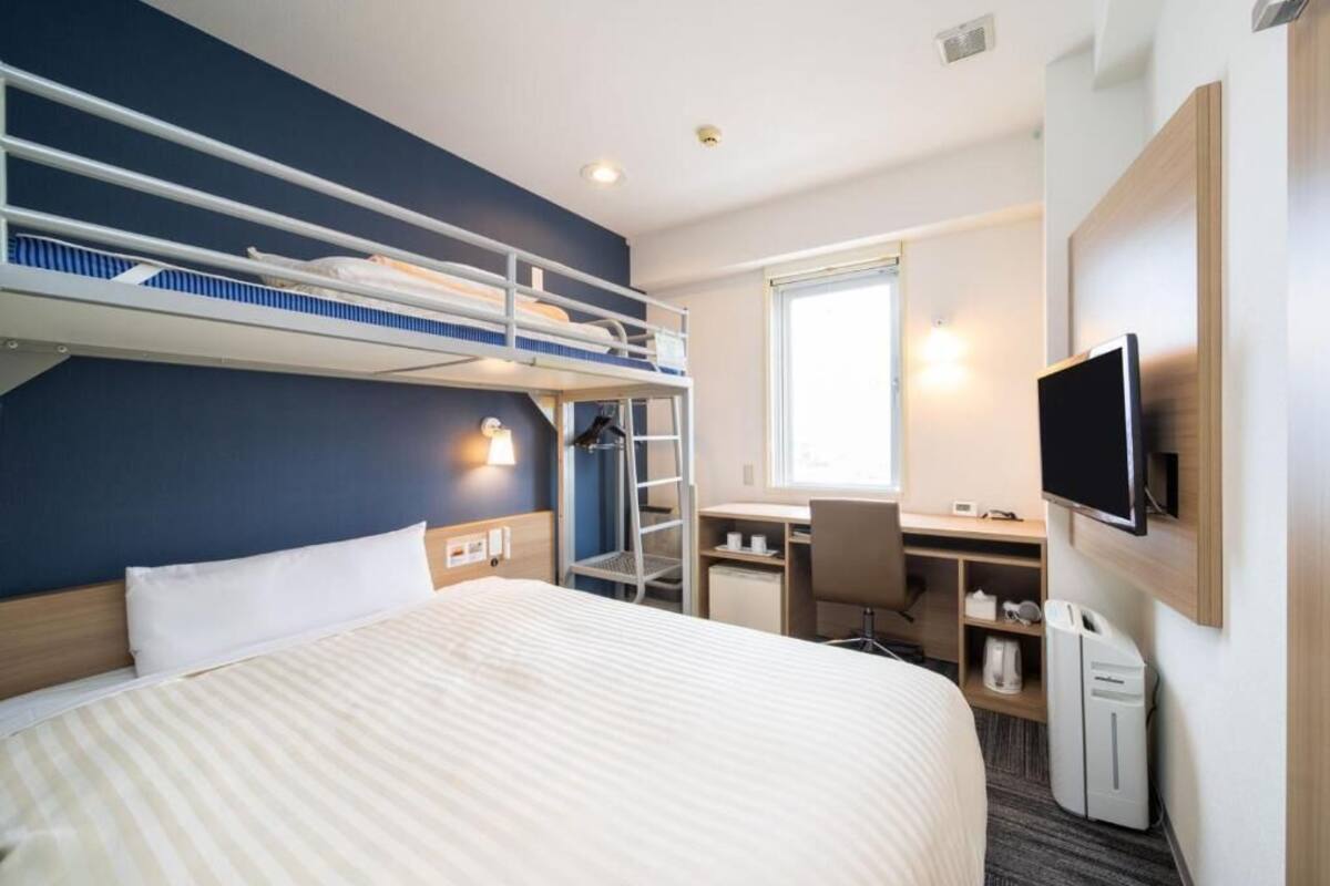 A hotel room with a bunk bed, a desk with a chair, a television, and a window.