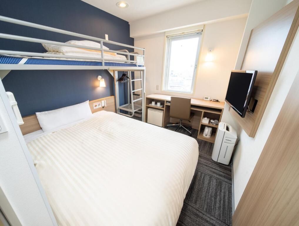 Une chambre d’hôtel avec un lit superposé, un bureau avec une chaise, une télévision et une fenêtre.