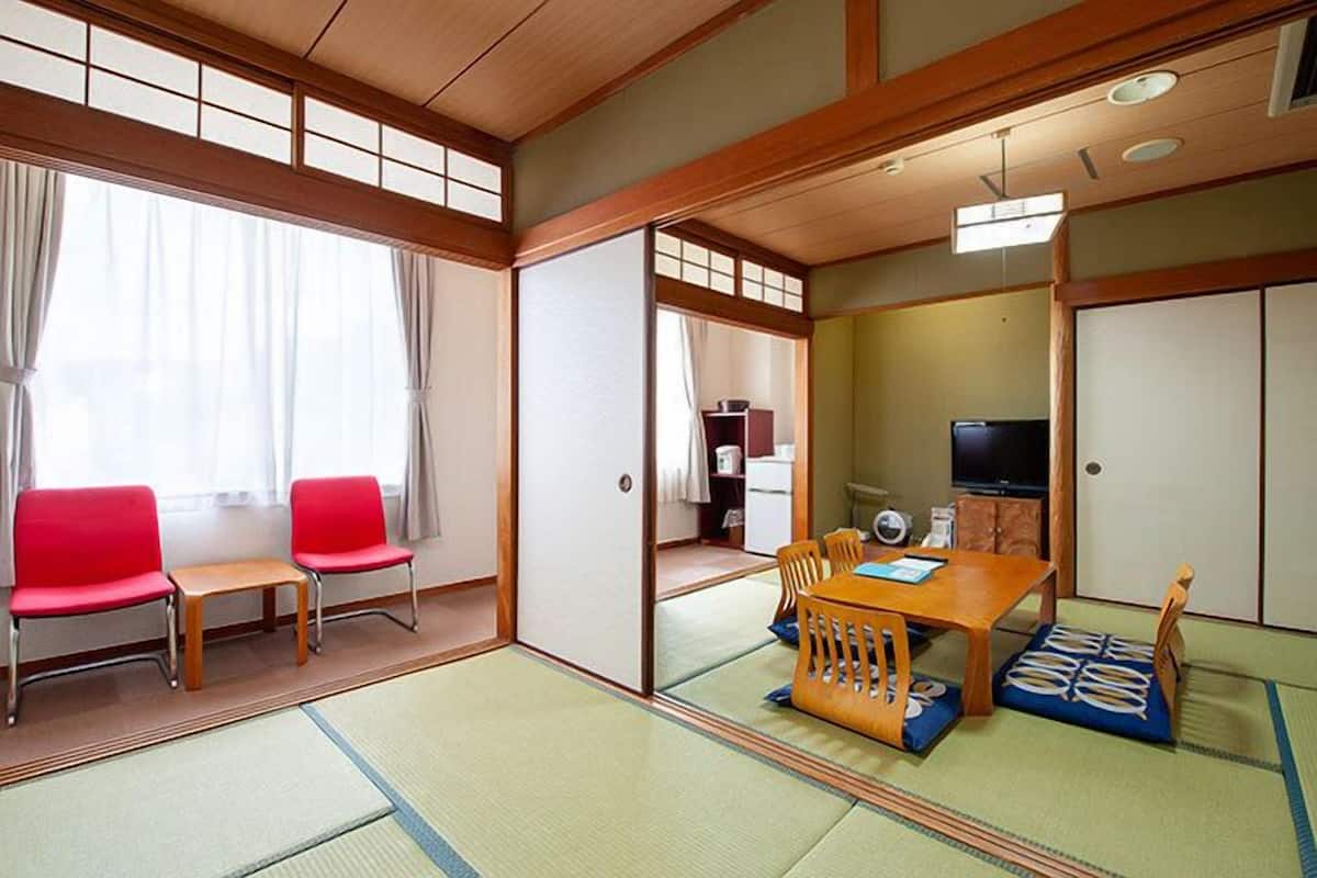Une pièce de style japonais traditionnel, avec un sol recouvert de tatamis, une petite table en bois, des chaises rouges et un téléviseur.