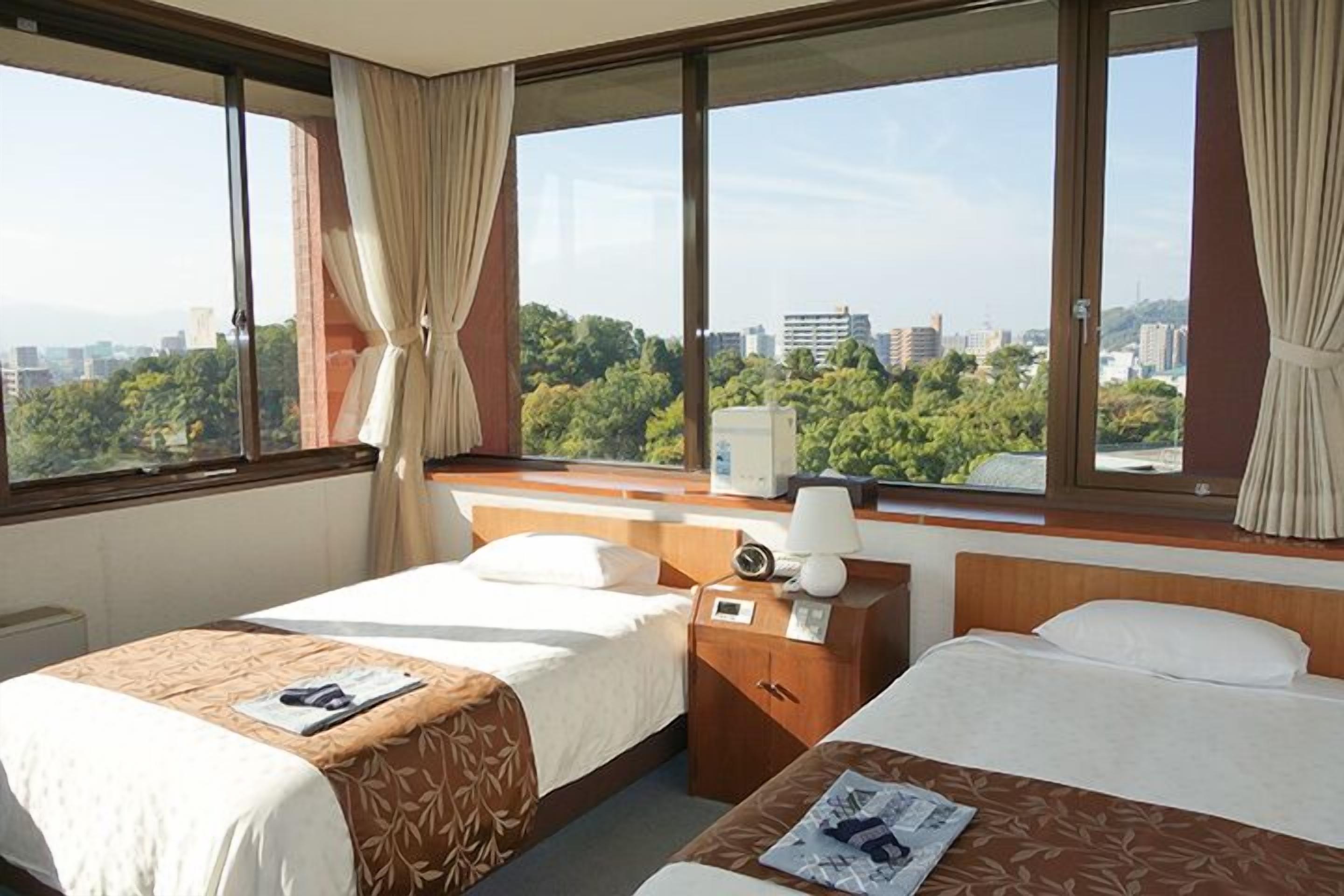 A hotel room with two beds, a window with curtains, a bedside table with a lamp, and a view of a cityscape.