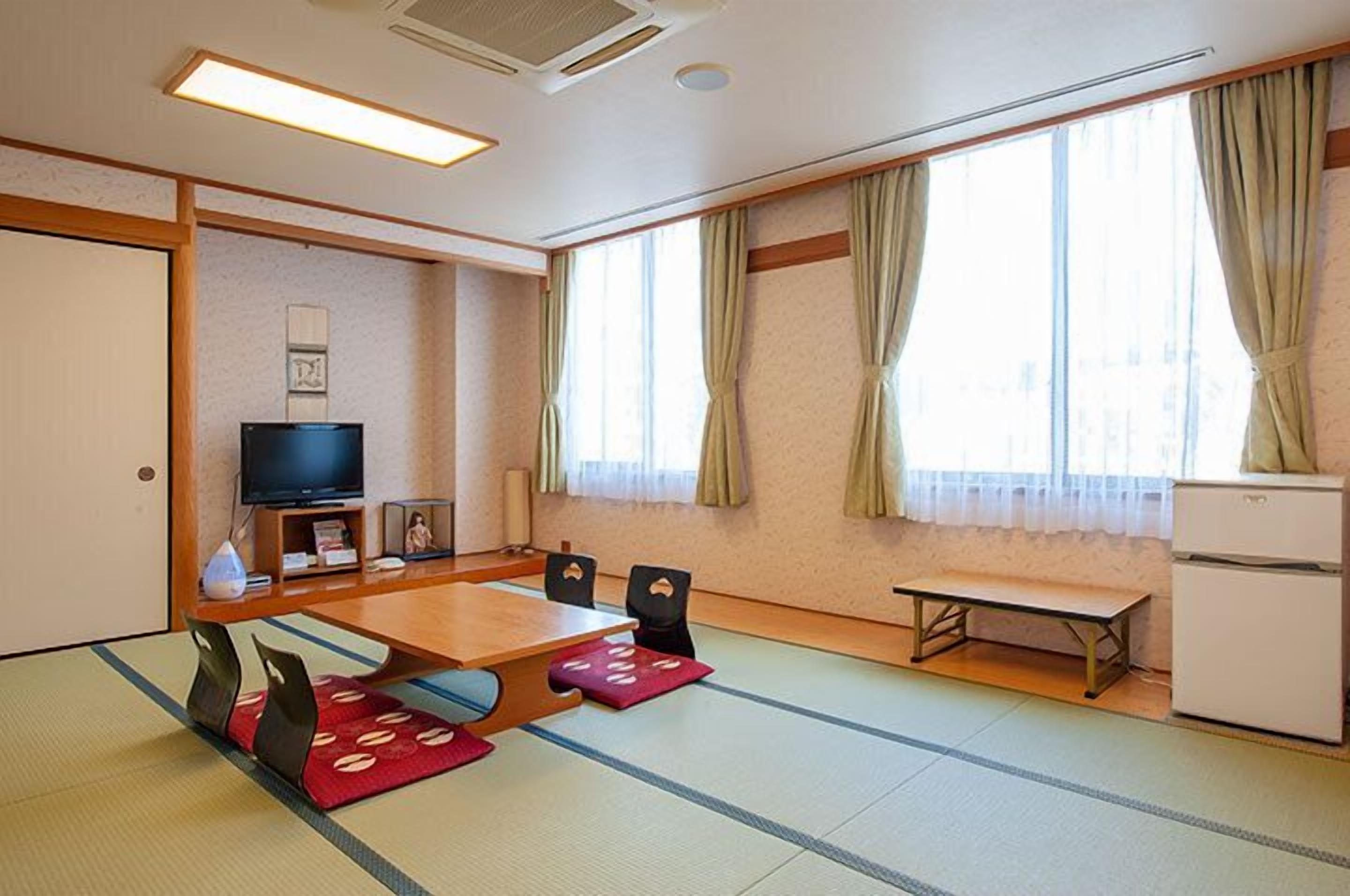 A room with tatami flooring, a wooden table, chairs, a television, a small refrigerator, and a window with curtains.