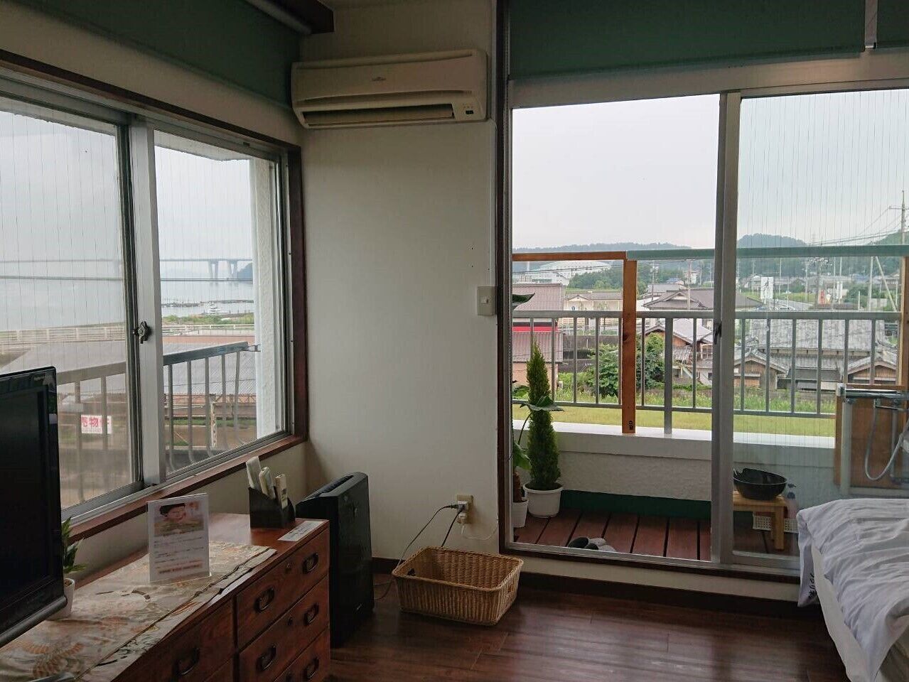 A room with a large window, a bed, a desk, and a small balcony with a potted plant.