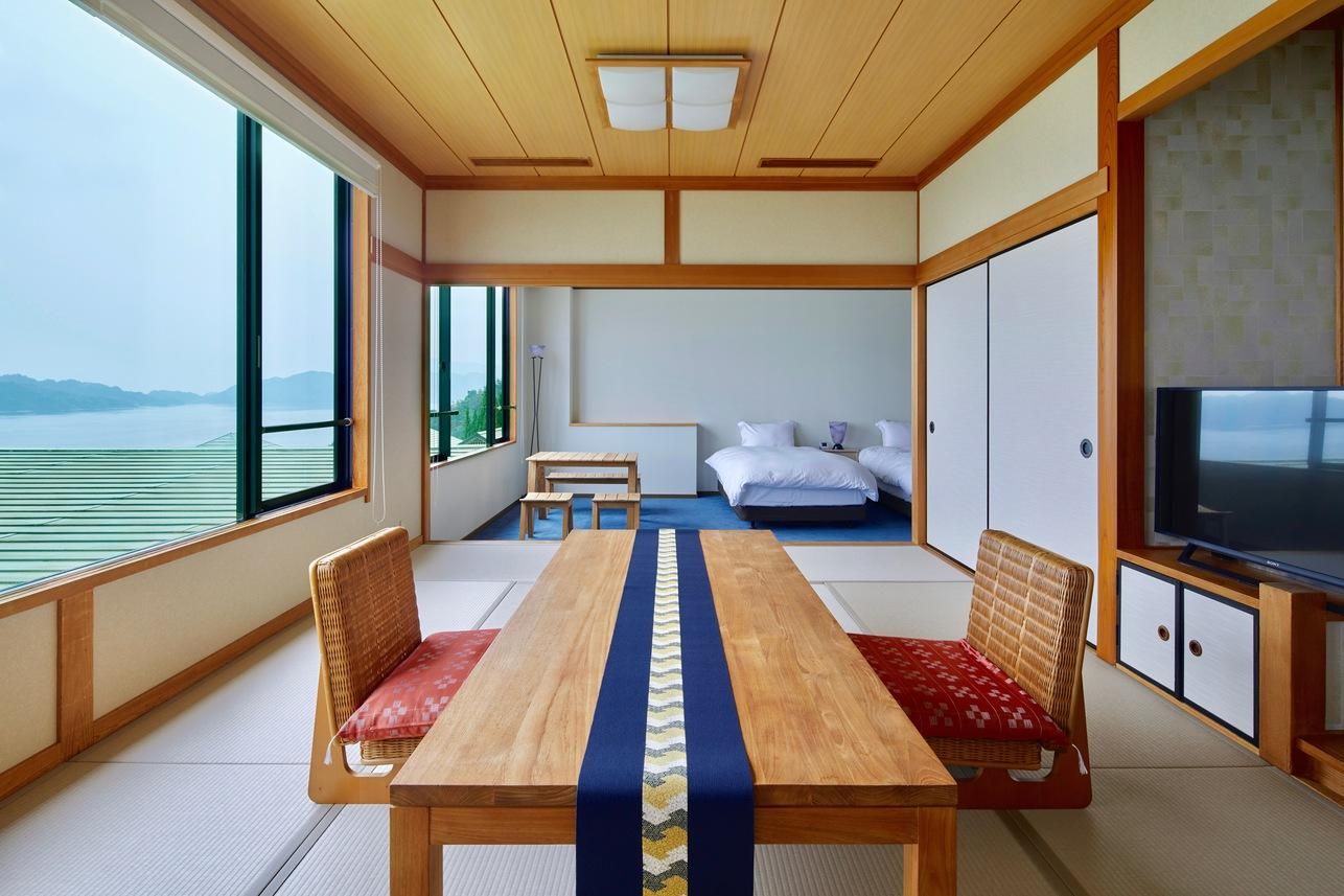 Quarto com mesa de madeira, duas cadeiras, uma cama, televisão e vista para o mar.