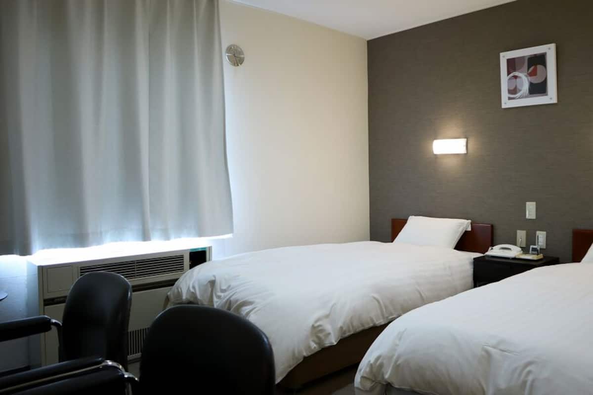 A hotel room with two beds, a desk, a chair, a window with curtains, and a wall-mounted clock.