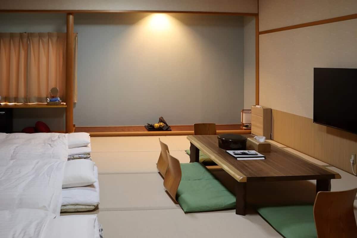 A room with a bed, a wooden table, a television, and a seating area with cushions.
