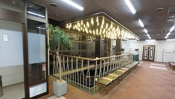 A hotel lobby with a long reception desk, a seating area with chairs, a plant, and a staircase leading to an upper level.