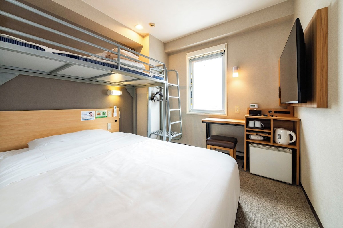 A hotel room with a bunk bed, a desk with a chair, a small kitchenette, and a television.