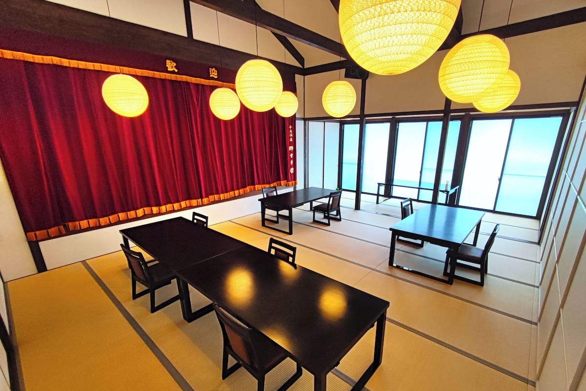 Een vergaderruimte met tatami matten, een podium met rode gordijnen en grote ramen.