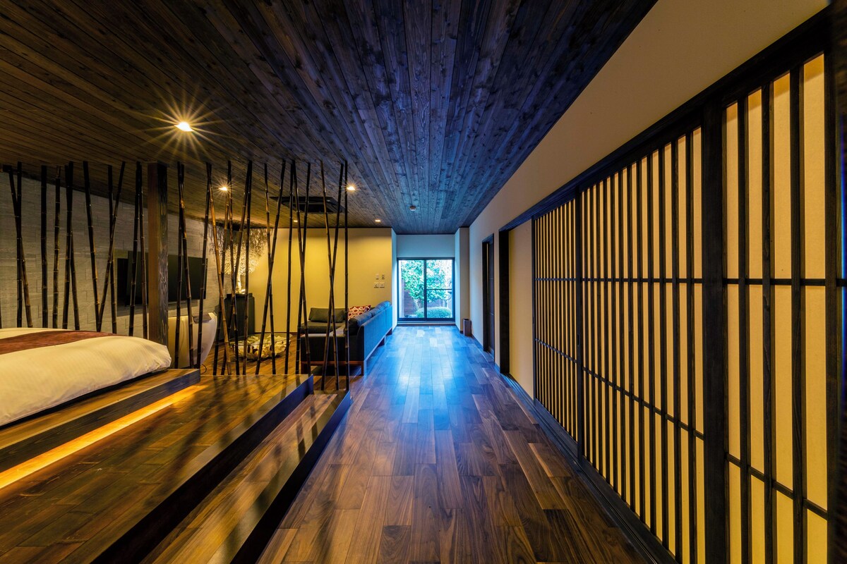 A modern hotel room with a wooden floor, a bed, a seating area, and a view of the outdoors.