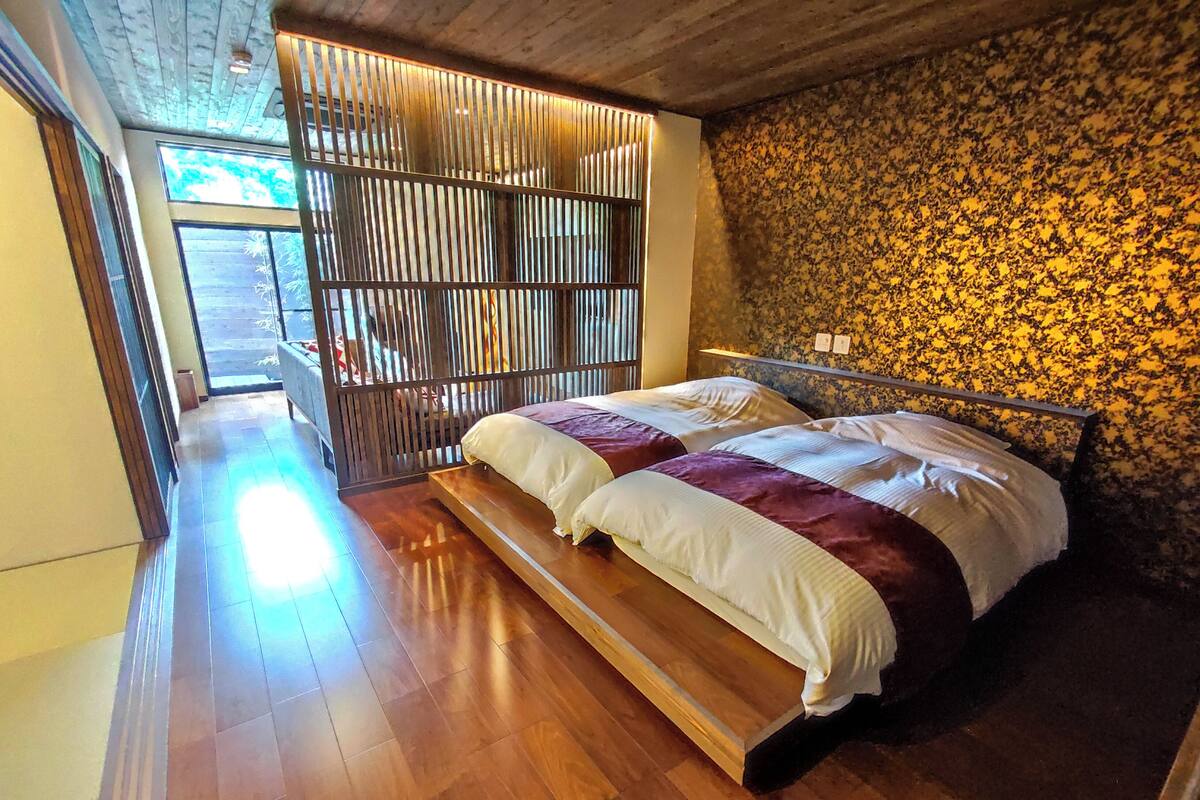 Une chambre avec un plancher en bois, un lit avec une literie blanche et violette, une tête de lit en bois et une fenêtre avec des stores.