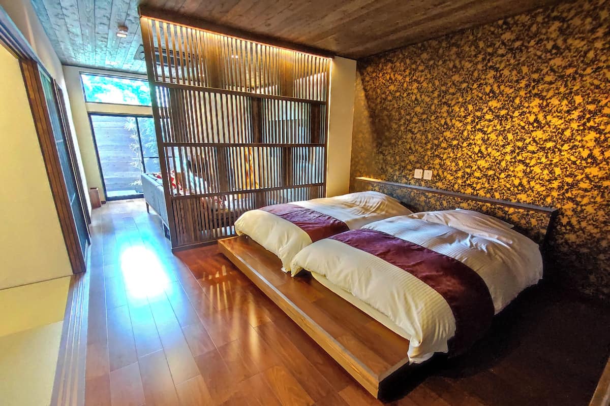 Een slaapkamer met een houten vloer, een bed met wit en paars beddengoed, een houten hoofdbord en een raam met jaloezieën.